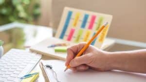 Teacher writing down notes on a clipboard while preparing a school project using colorful sticky notes and a pencil, with a computer keyboard and a plant in the background